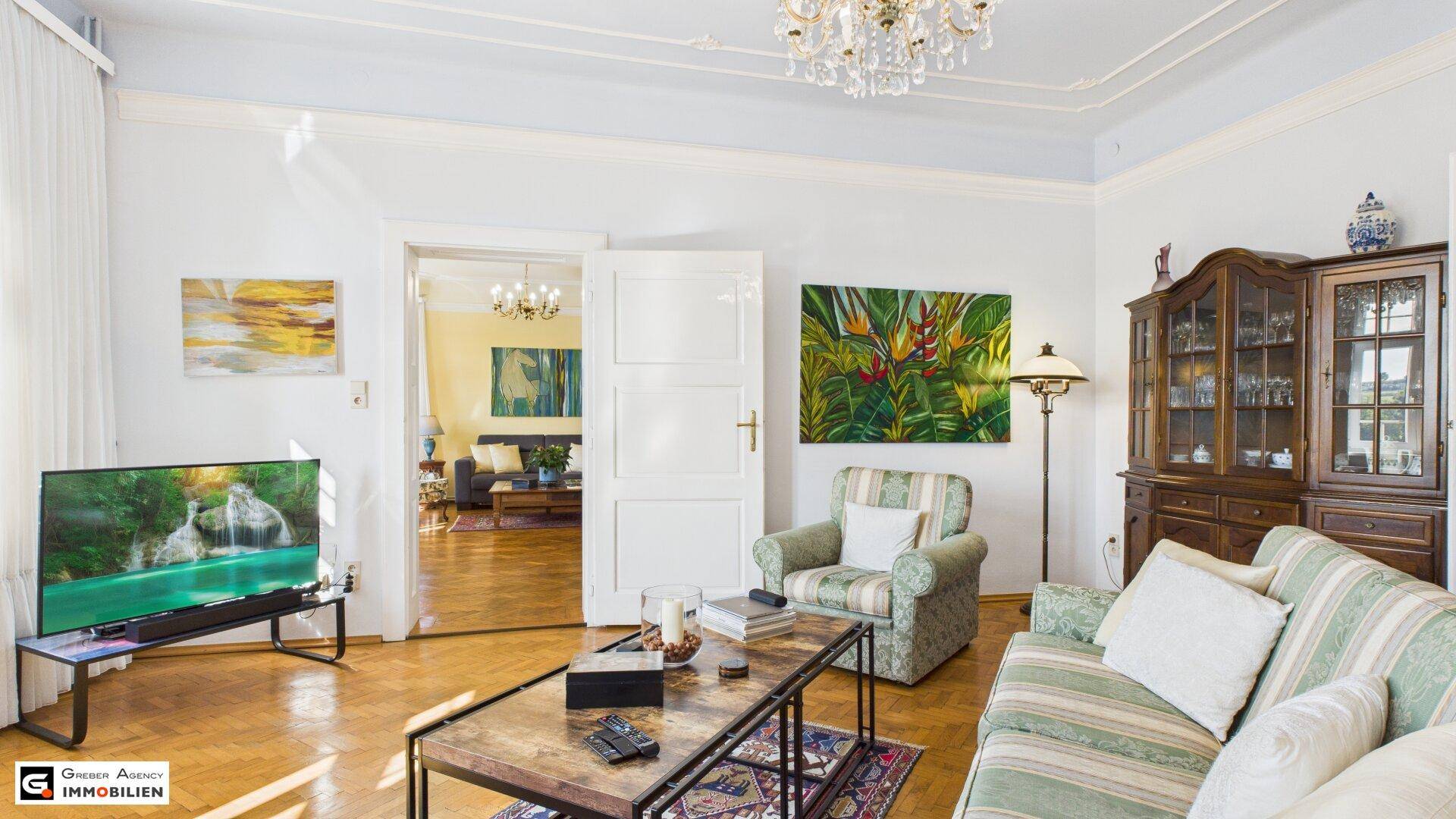Geräumiges Wohnzimmer mit Parkettboden, Stuckdecke und Blick in den angrenzenden Raum.