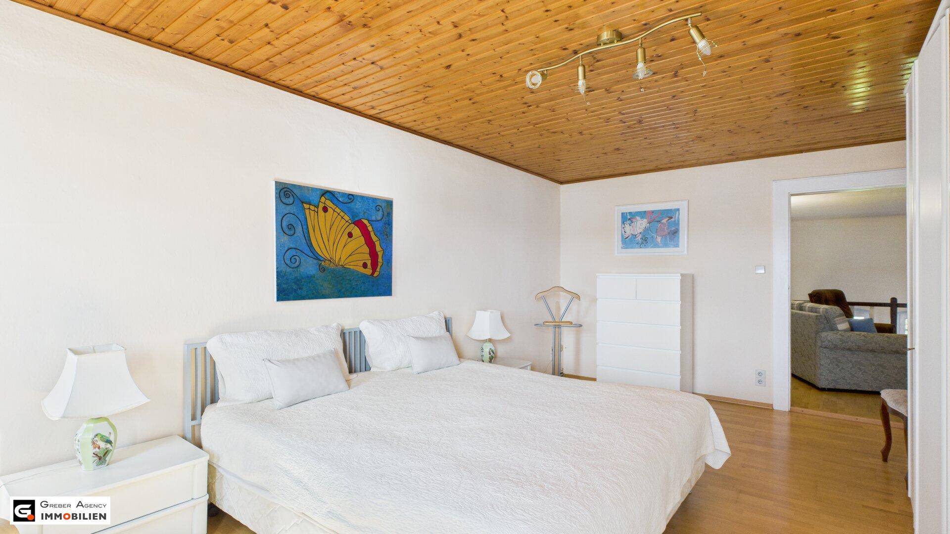Schlafzimmer mit Holzbalkendecke, Doppelbett und Blick in den angrenzenden Wohnbereich.