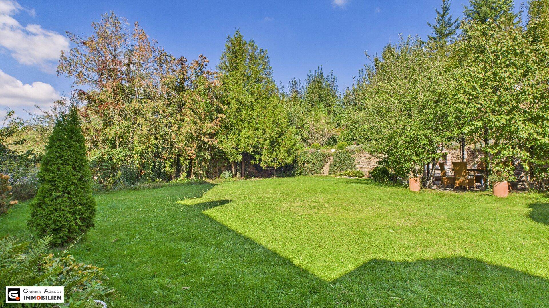 Gepflegter Garten mit Rasenfläche, Bäumen und einer gemütlichen Sitzecke im Hintergrund.