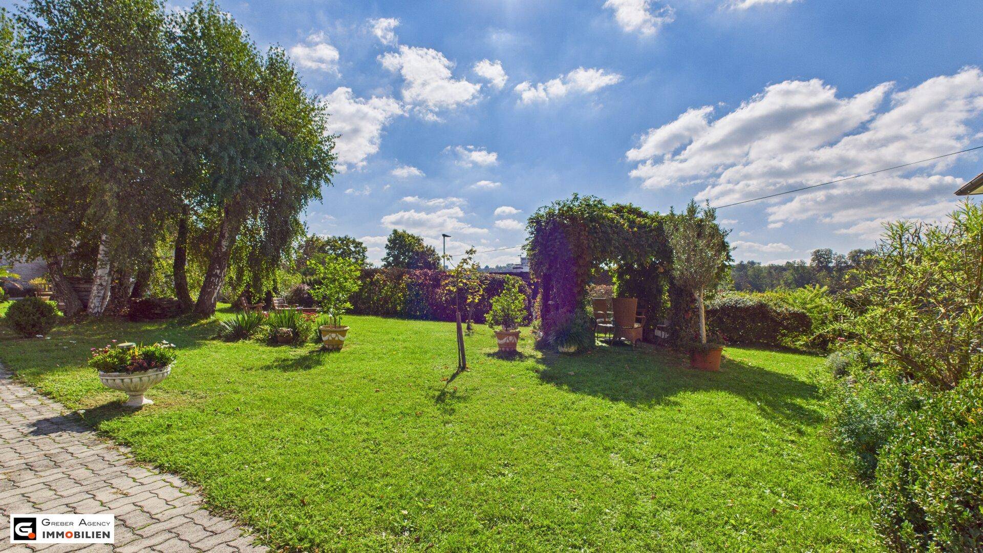 Weitläufiger Garten mit Rasen, Bäumen und einem gepflasterten Weg unter blauem Himmel.