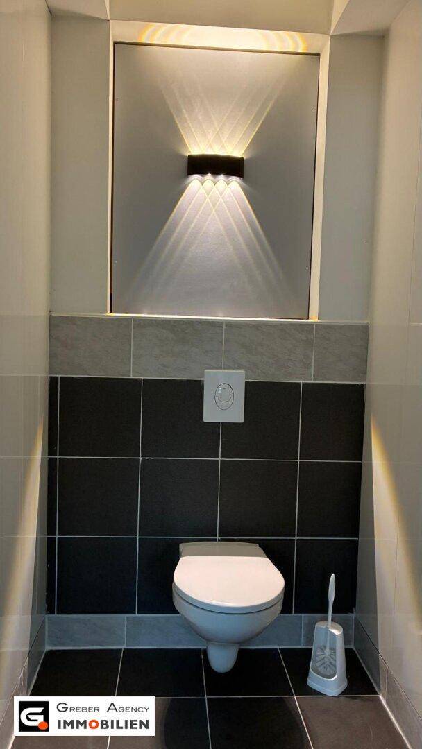 Modernes Gäste-WC mit Wand-WC, dunklen Fliesen und indirekter Beleuchtung.