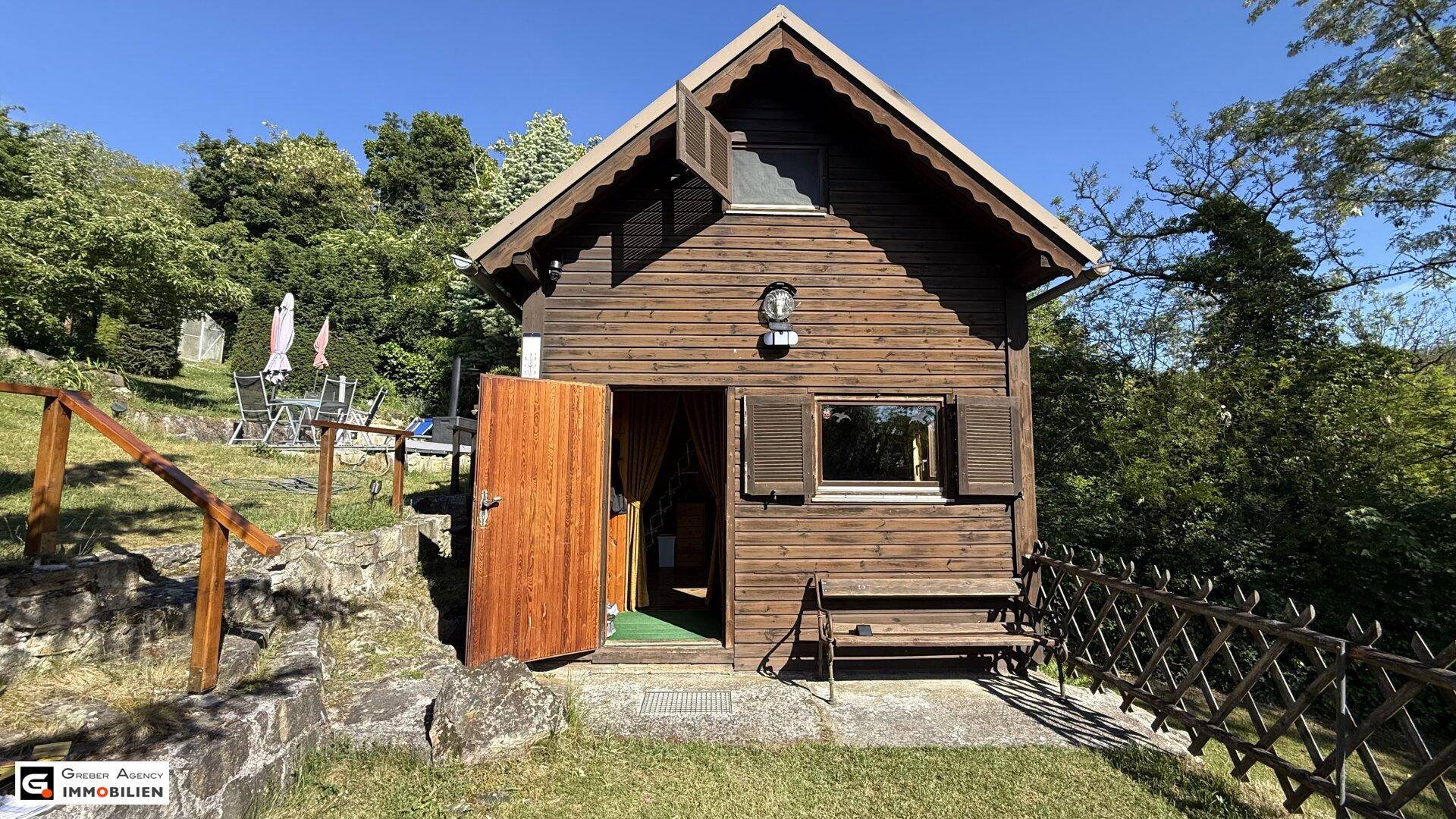 Charmantes Holzhaus mit Satteldach und offener Tür, umgeben von üppiger Vegetation.
