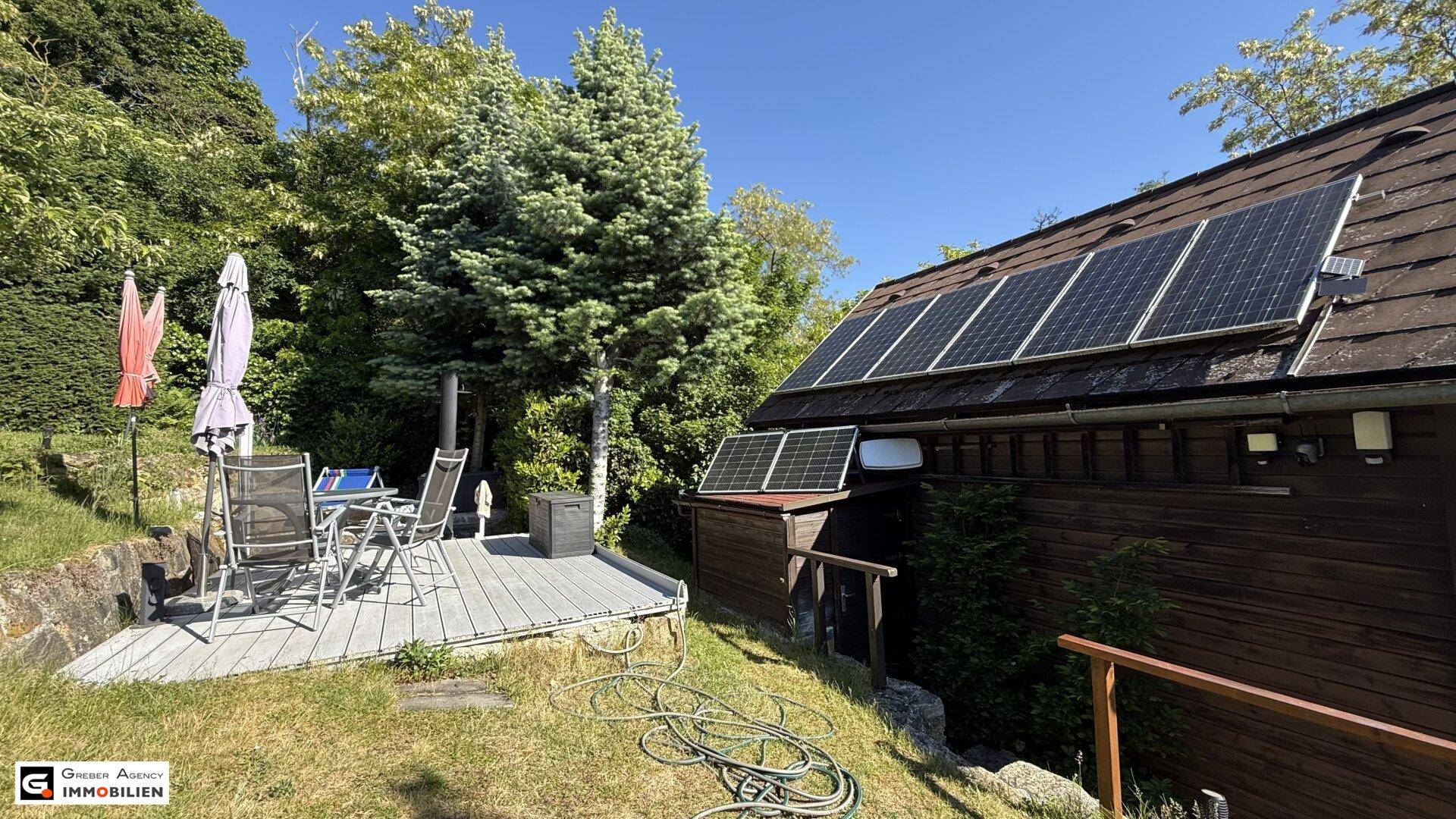 Terrasse mit Gartenmöbeln und Sonnenschirmen, daneben Solarpaneele auf dem Dach des Holzhauses.