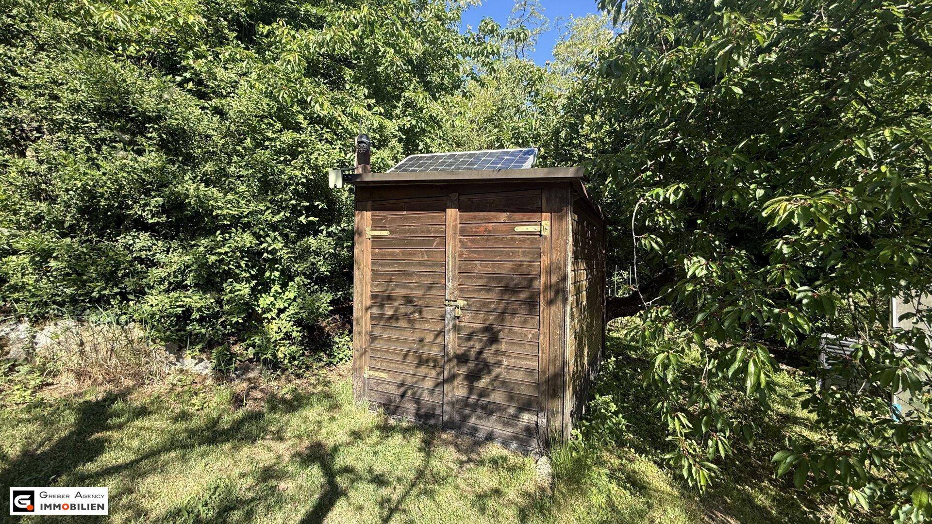Kleiner Holzschuppen im Garten mit Solarpaneel auf dem Dach, umgeben von Bäumen.