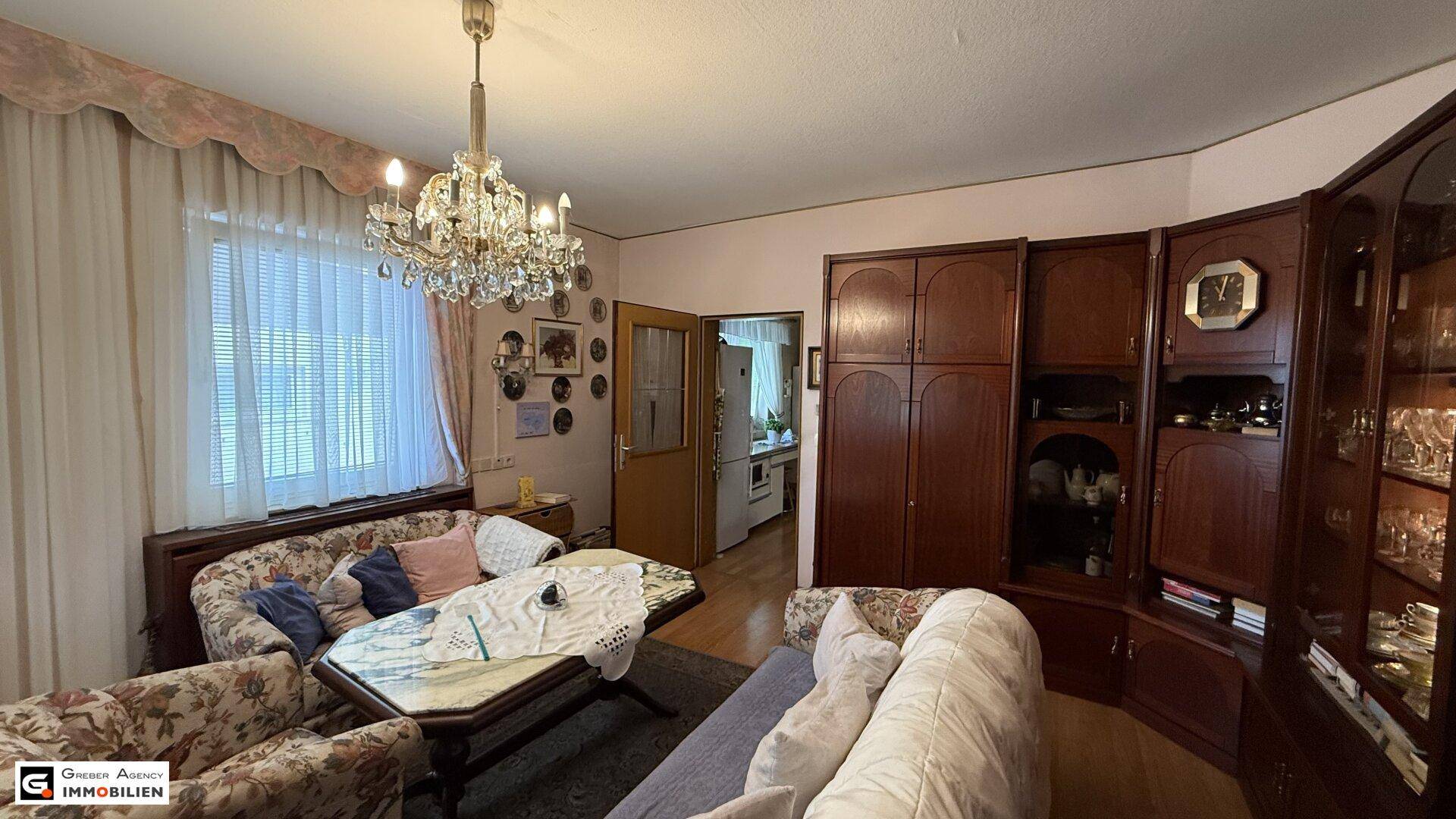 Geräumiges Wohnzimmer mit dunklen Holzmöbeln, Kronleuchter und Zugang zur Küche, gut erhalten.