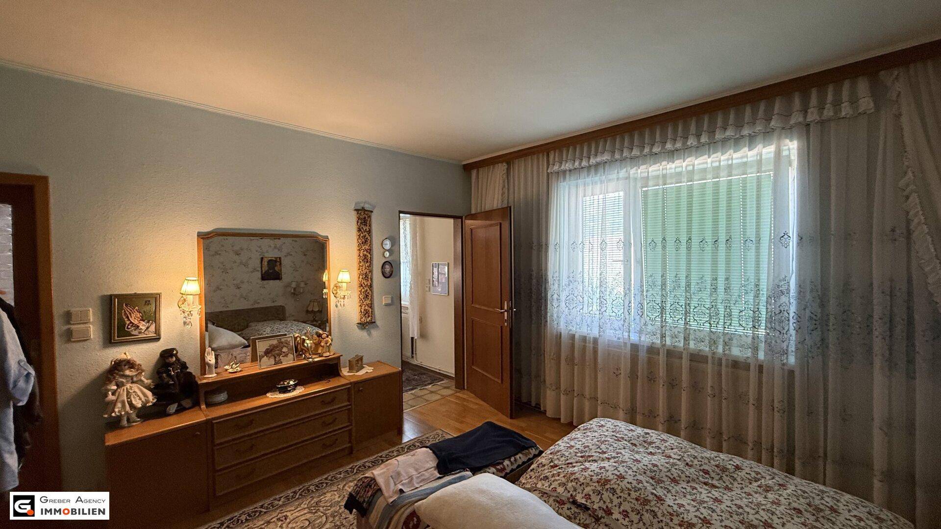 Helles Schlafzimmer mit klassischer Einrichtung und Blick ins Grüne durch das große Fenster.