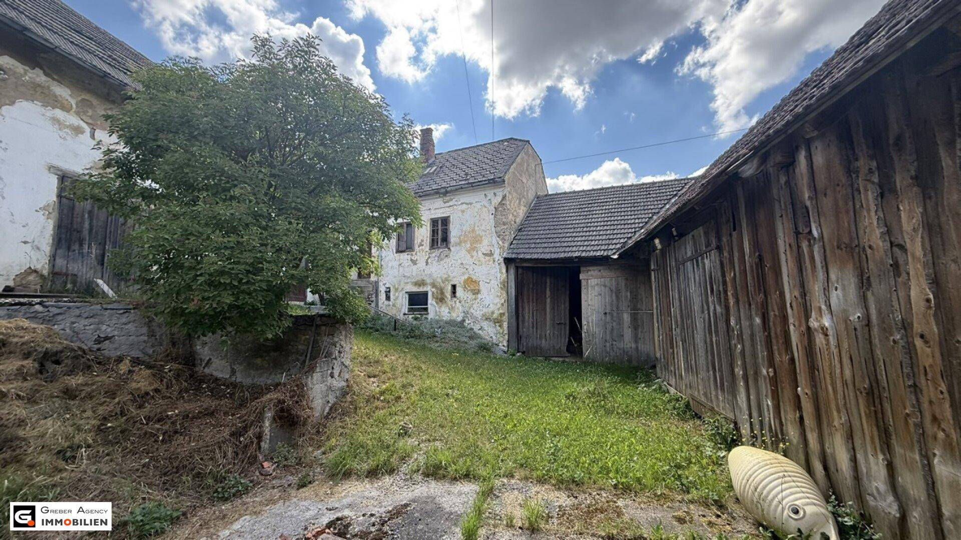 Verwilderter Gartenbereich mit alten Gebäuden, darunter ein baufälliges Wohnhaus und eine Holzscheune.