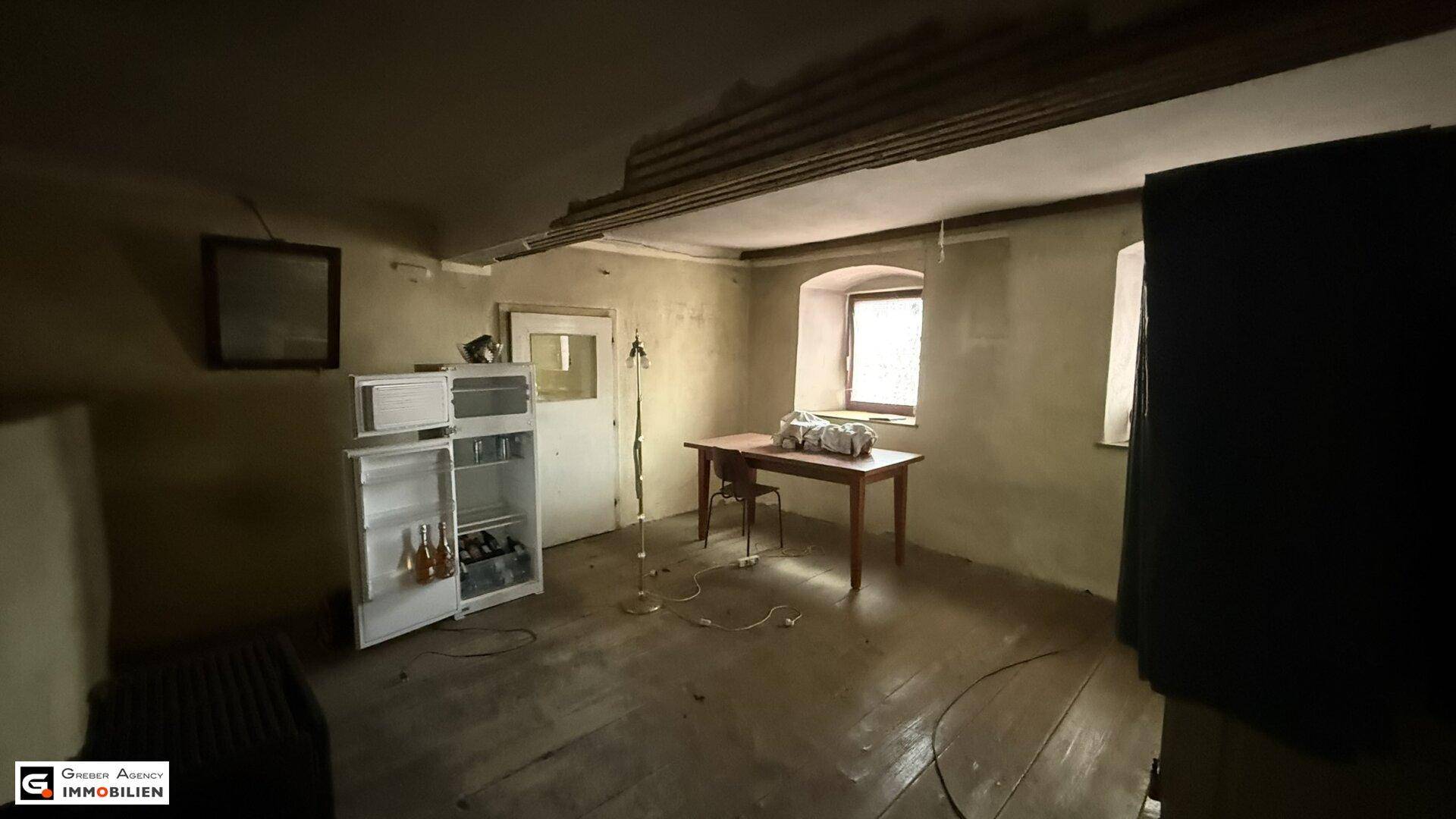 Dunkler Innenraum mit Holzboden, einem Tisch, Kühlschrank und kleinen Fenstern, in schlechtem Zustand.