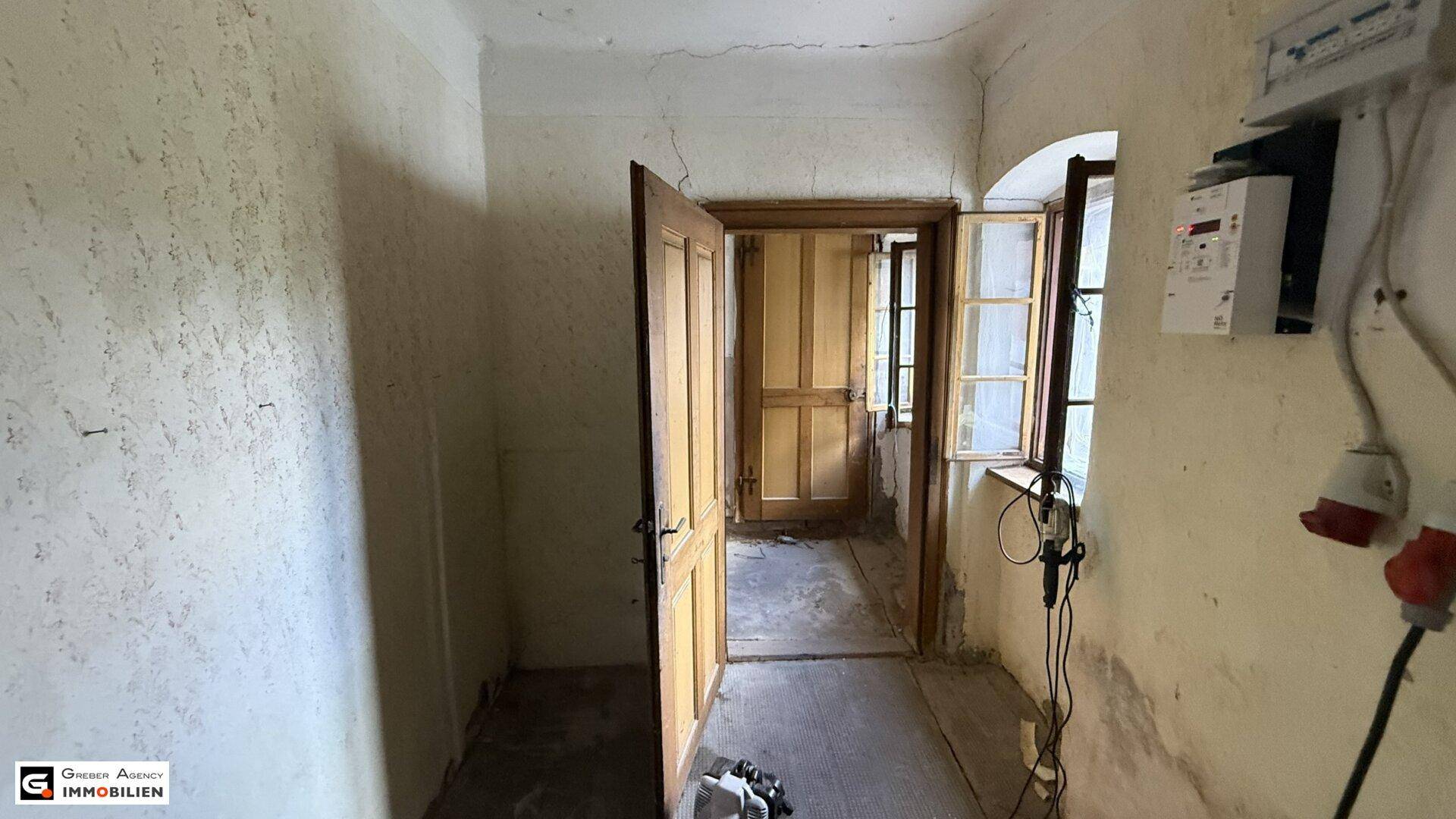 Alter Flur mit Holztüren, einem Fenster und sichtbaren elektrischen Installationen.