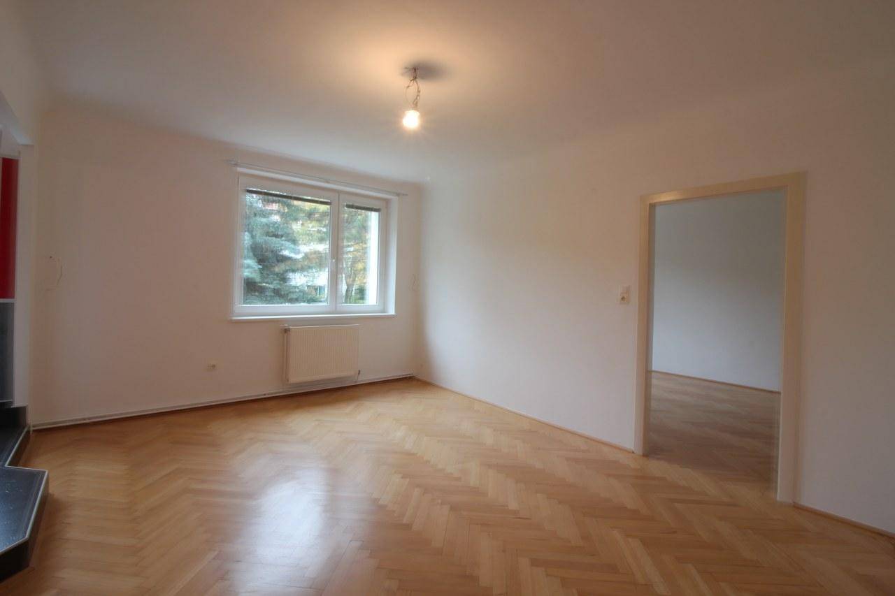 Heller Wohnraum mit Fischgrätparkett, großem Fenster und Zugang zu einem weiteren Zimmer.