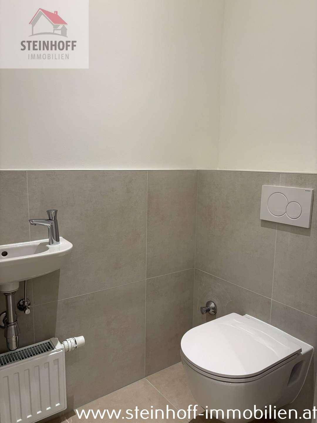 Modernes Gäste-WC mit Wand-WC, kleinem Waschbecken und hellen Fliesen an Wänden und Boden.