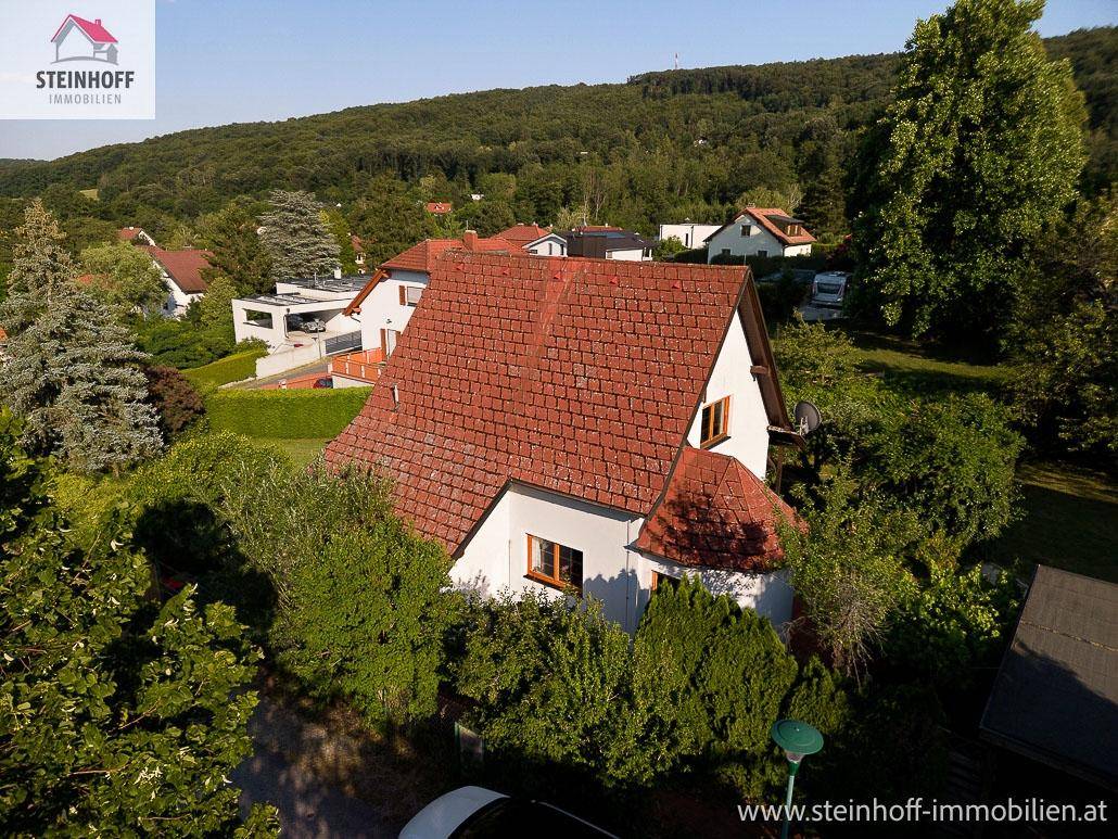 Ansicht des Hauses mit rotem Ziegeldach, umgeben von Bäumen und Hügeln unter blauem Himmel.