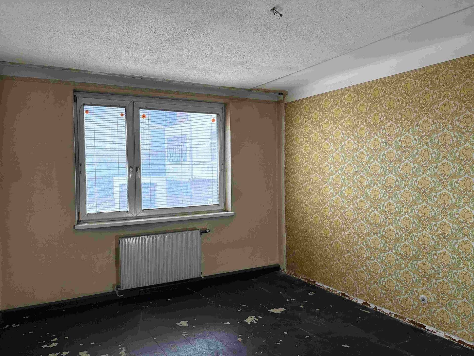 Leeres Zimmer mit dunklem, abgenutztem Boden, Heizkörper und gemusterter Tapete an einer Wand.
