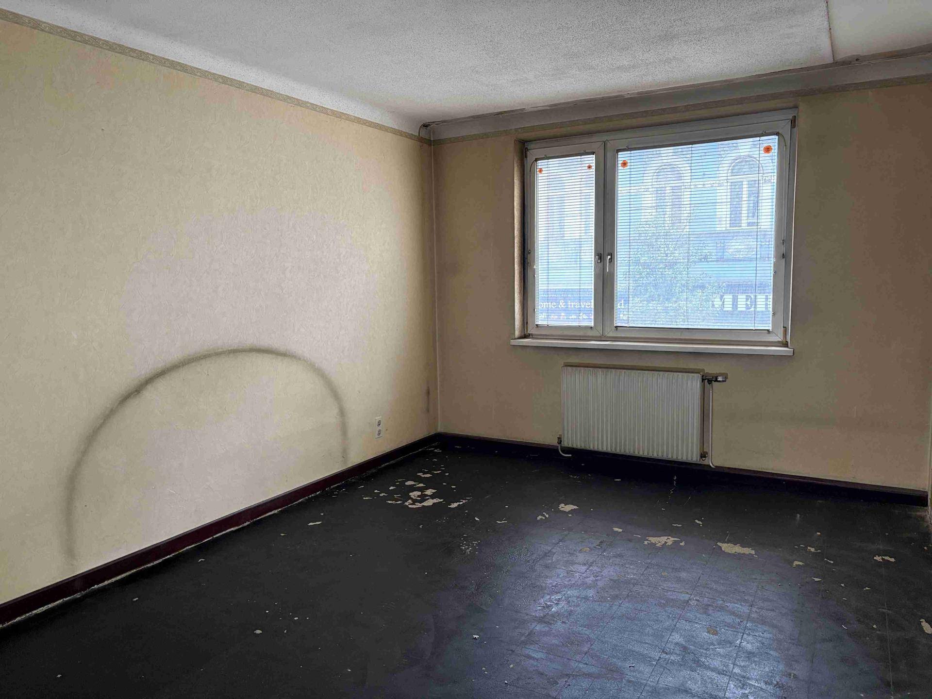 Leeres Zimmer mit heller Tapete und Fenster mit Jalousien, das auf ein Gebäude blickt.
