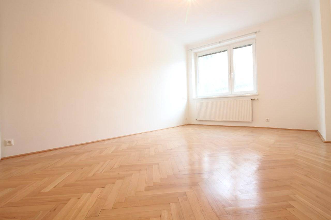 Leerer Raum mit Fischgrätparkett und großem Fenster, das viel Tageslicht hereinlässt.