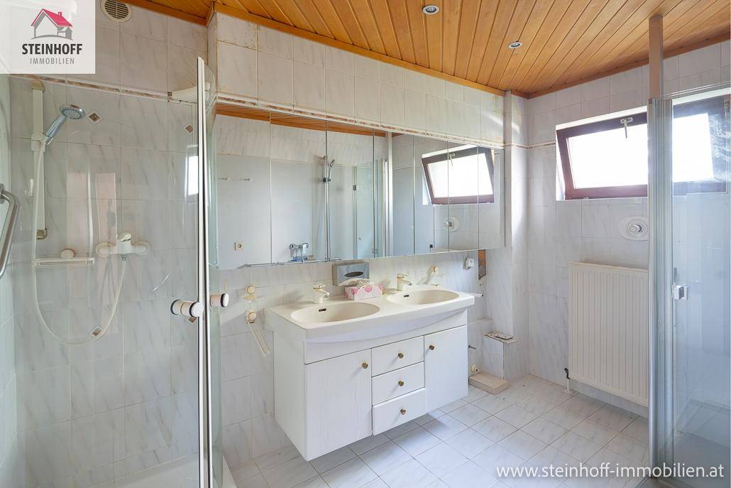 Helles Badezimmer mit Doppelwaschbecken, Duschkabinen, weißen Fliesen und Holzdecke.