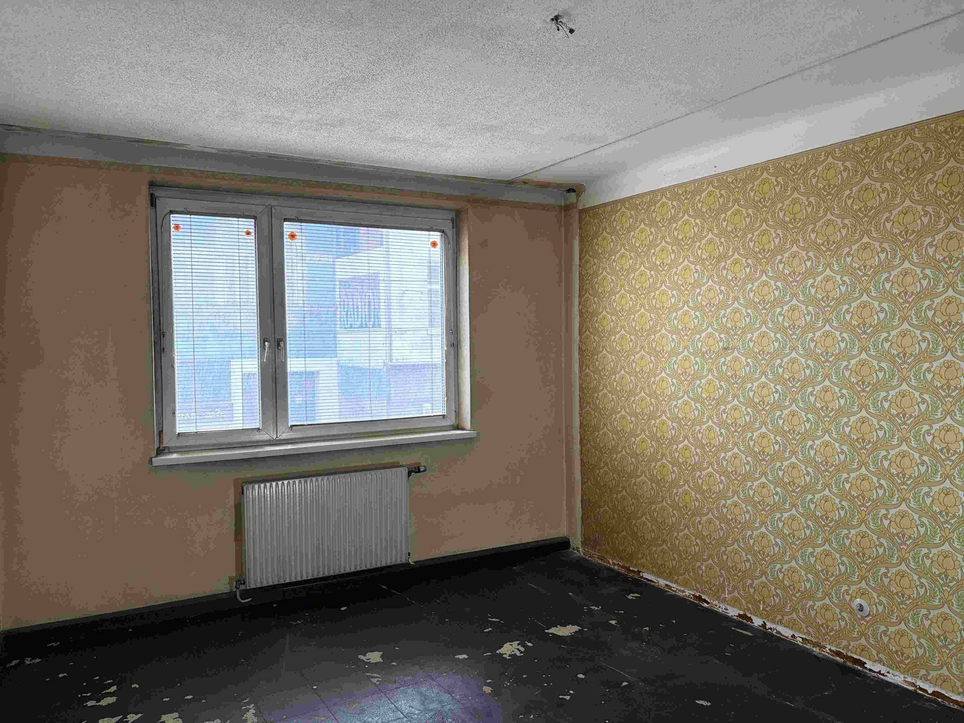 Leeres Zimmer mit gemusterter Tapete und Fenster mit Jalousien, das auf ein Gebäude blickt.