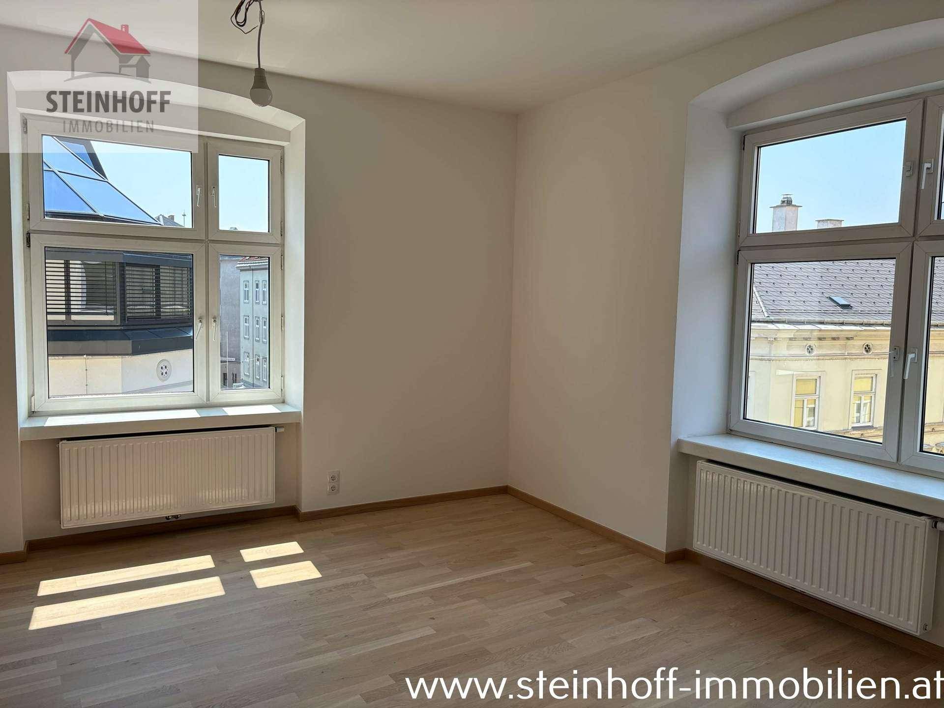Heller Raum mit Parkettboden und zwei Fenstern, die einen Blick auf die umliegenden Gebäude ermöglichen.
