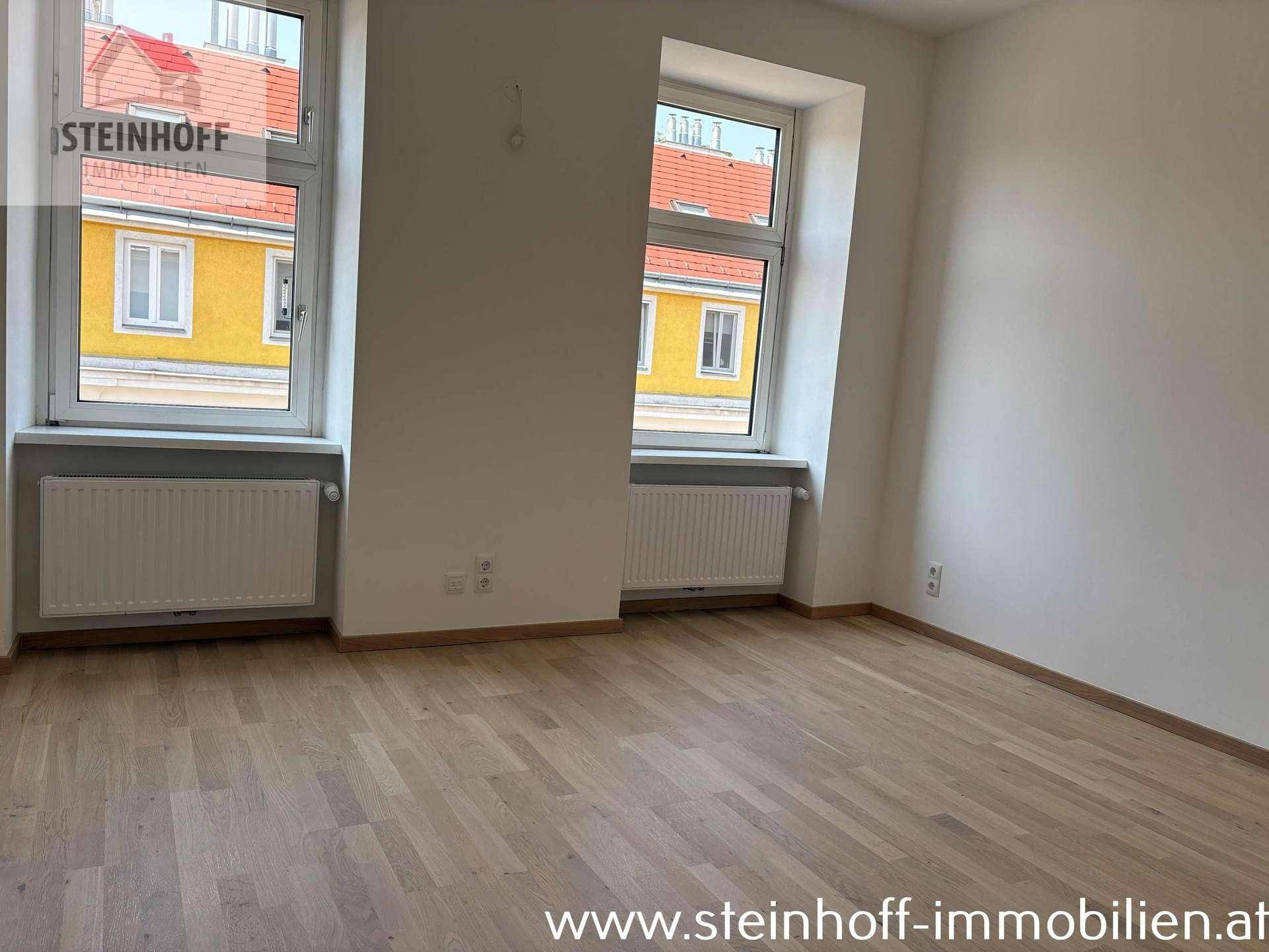 Heller Raum mit Parkettboden und zwei Fenstern, die einen Blick auf die gegenüberliegenden Häuser bieten.
