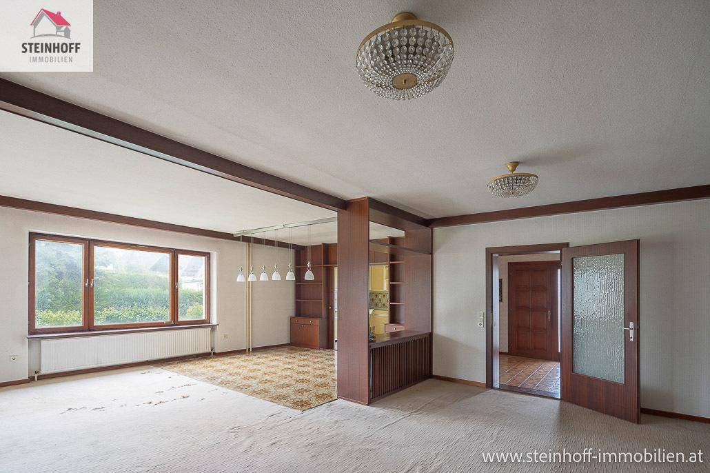 Unmöbliertes Wohnzimmer mit Teppichboden, Holzbalken und Blick in den Garten.