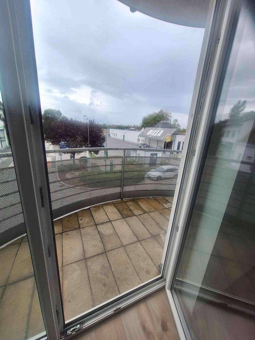 Blick durch die offene Balkontür auf den gefliesten Balkon und die belebte Straße mit Geschäften.