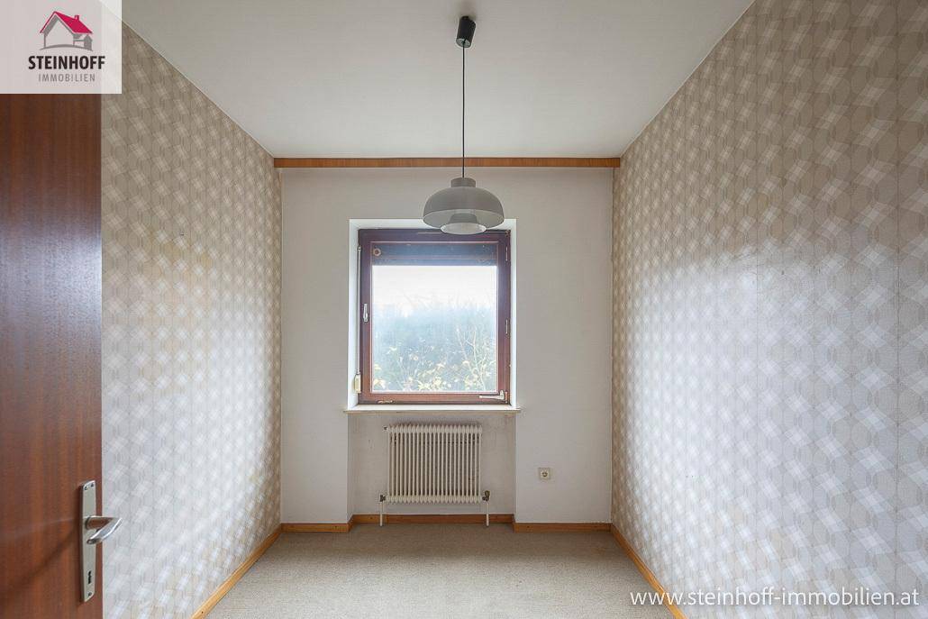 Kleines Zimmer mit gemusterter Tapete, Teppichboden und einem Fenster.