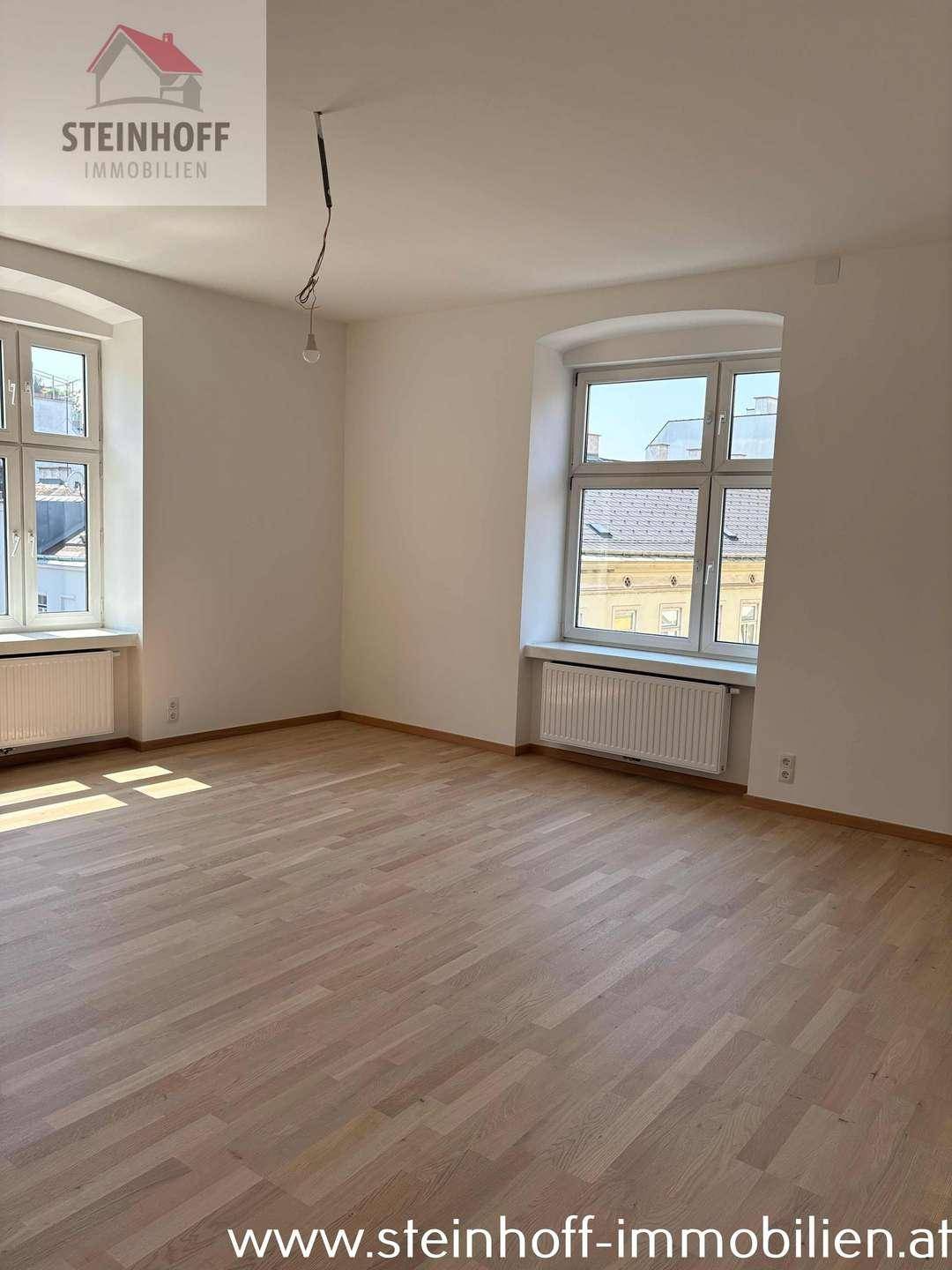 Lichtdurchfluteter Raum mit Parkettboden und zwei Fenstern, die einen Blick nach draußen bieten.