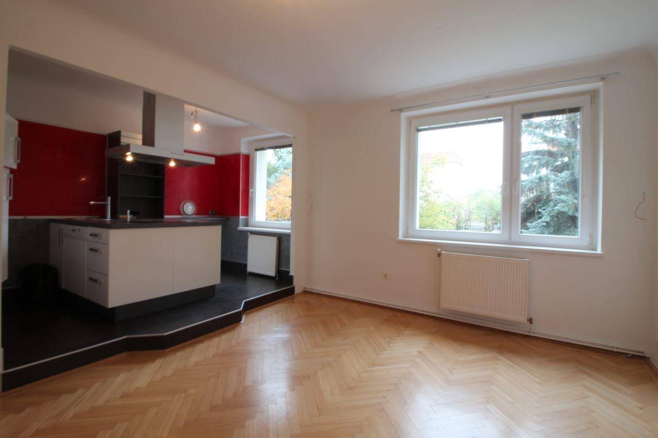 Heller Wohnbereich mit offener Küche, Fischgrätparkett und Fenster mit Blick auf Bäume.