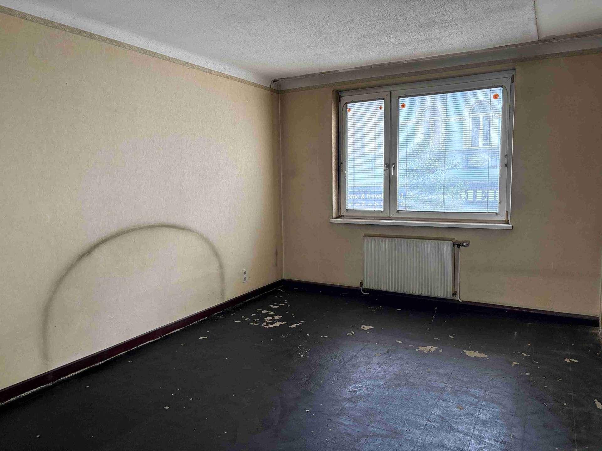 Leeres Zimmer mit dunklem, abgenutztem Boden, Heizkörper und Fenster mit Jalousien.