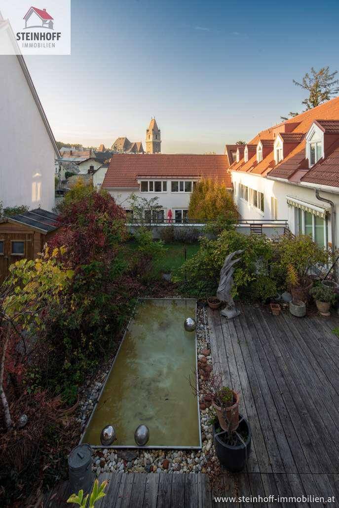 Gepflegter Garten mit Holzterrasse, Wasserbecken und üppiger Bepflanzung, umgeben von Häusern.