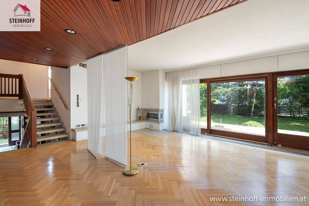 Großzügiges Wohnzimmer mit Parkettboden, Holzdecke, Kamin und Zugang zum Garten durch große Schiebetüren.