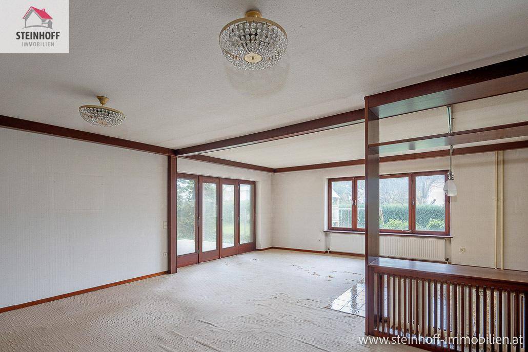 Unmöbliertes Wohnzimmer mit Teppichboden, Holzbalken und Zugang zum Garten.