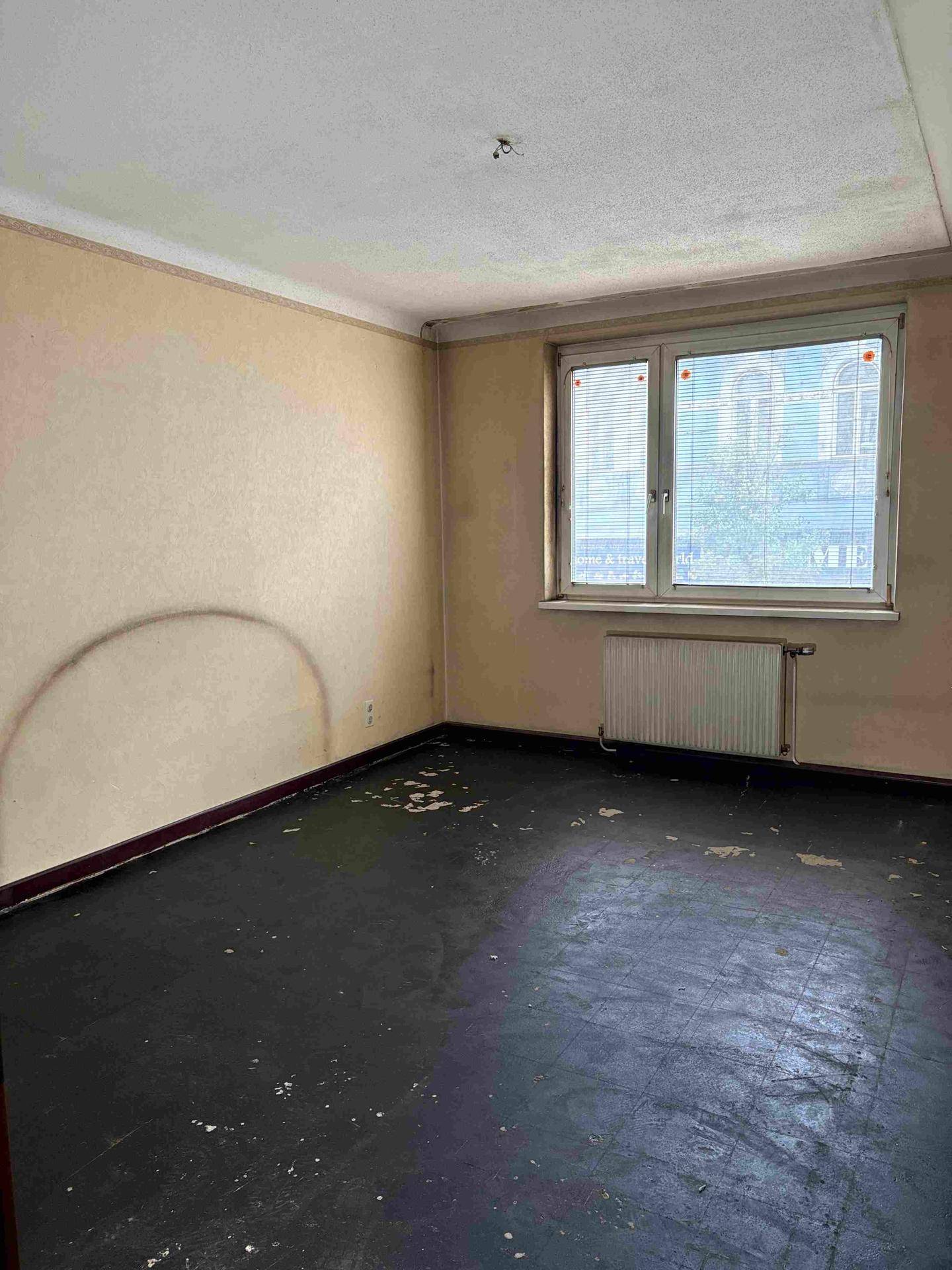 Leeres Zimmer mit heller Tapete und Fenster mit Jalousien, das auf ein Gebäude blickt.