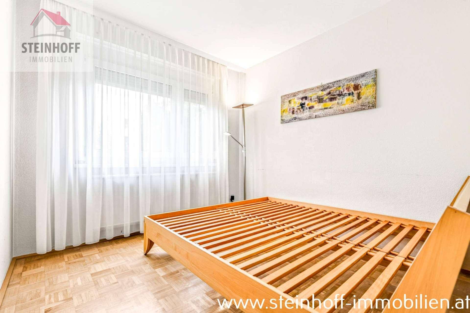 Helles Schlafzimmer mit Holzbettrahmen, Parkettboden und großem Fenster mit Vorhängen.