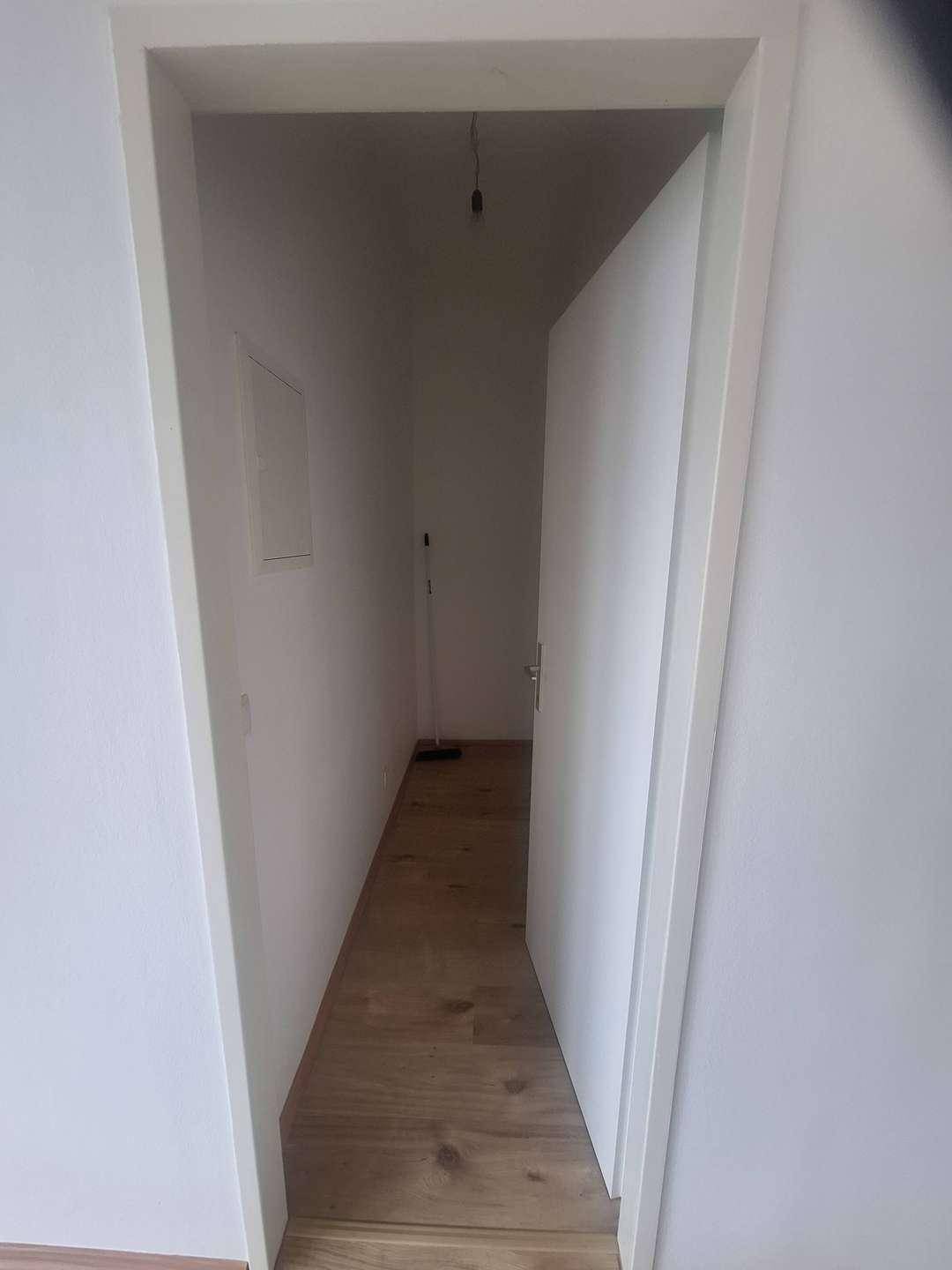 Heller, schmaler Flur mit Laminatboden und weißen Wänden, eine offene Tür führt in den nächsten Raum.