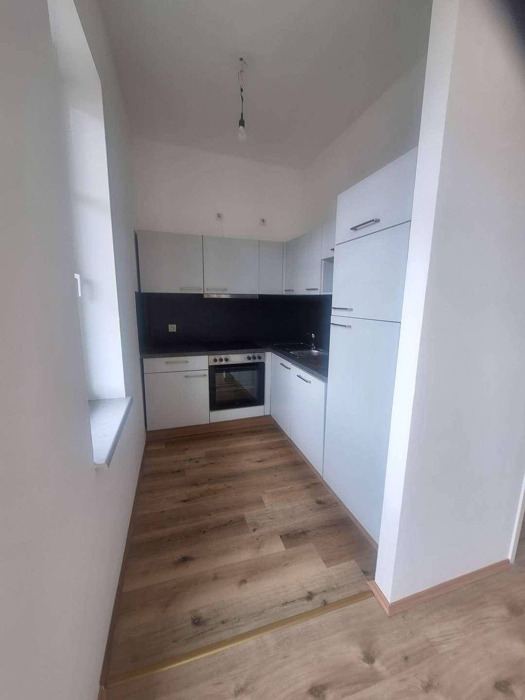 Schmale, moderne Küche mit weißen Schränken, dunkler Arbeitsplatte und hellem Laminatboden, inklusive Backofen.