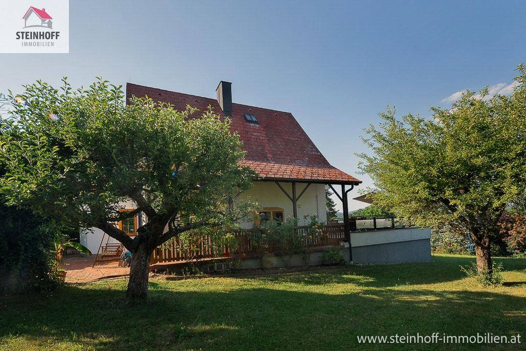 Haus mit Garten und Terrasse, umgeben von Bäumen unter blauem Himmel.