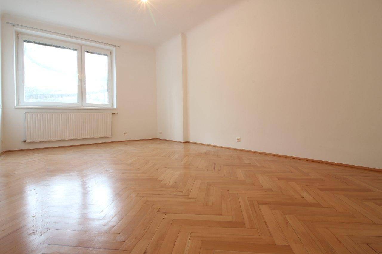 Unmöblierter Raum mit Fischgrätparkett und großem Fenster, das den Raum erhellt.