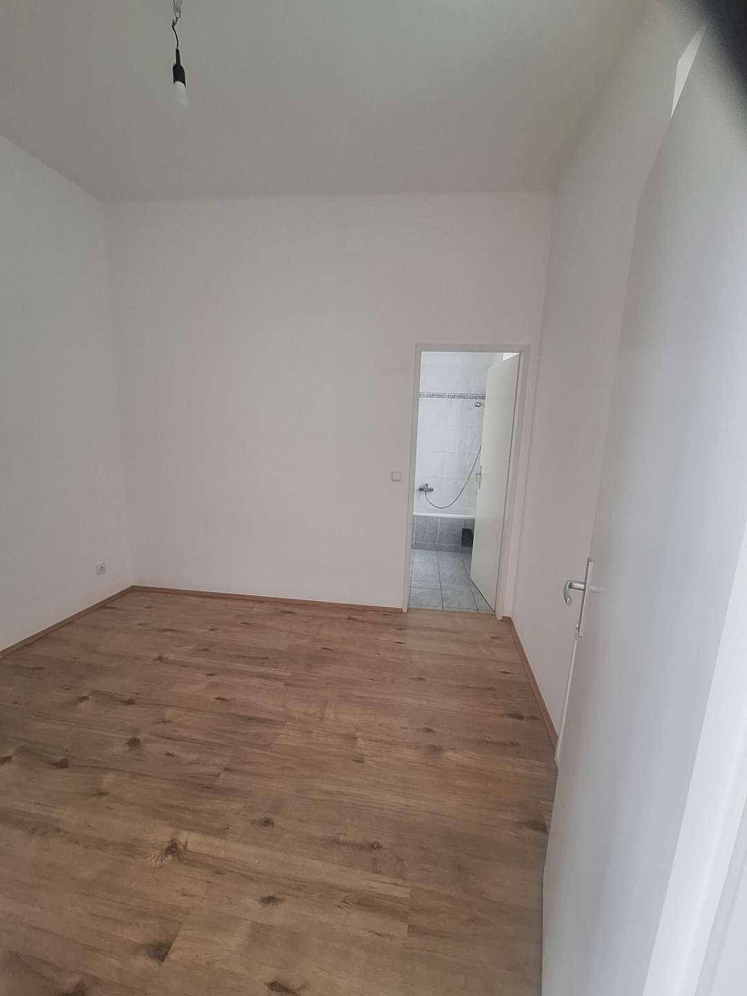 Leeres Zimmer mit hellem Laminatboden und weißen Wänden, mit Zugang zu einem gefliesten Badezimmer.