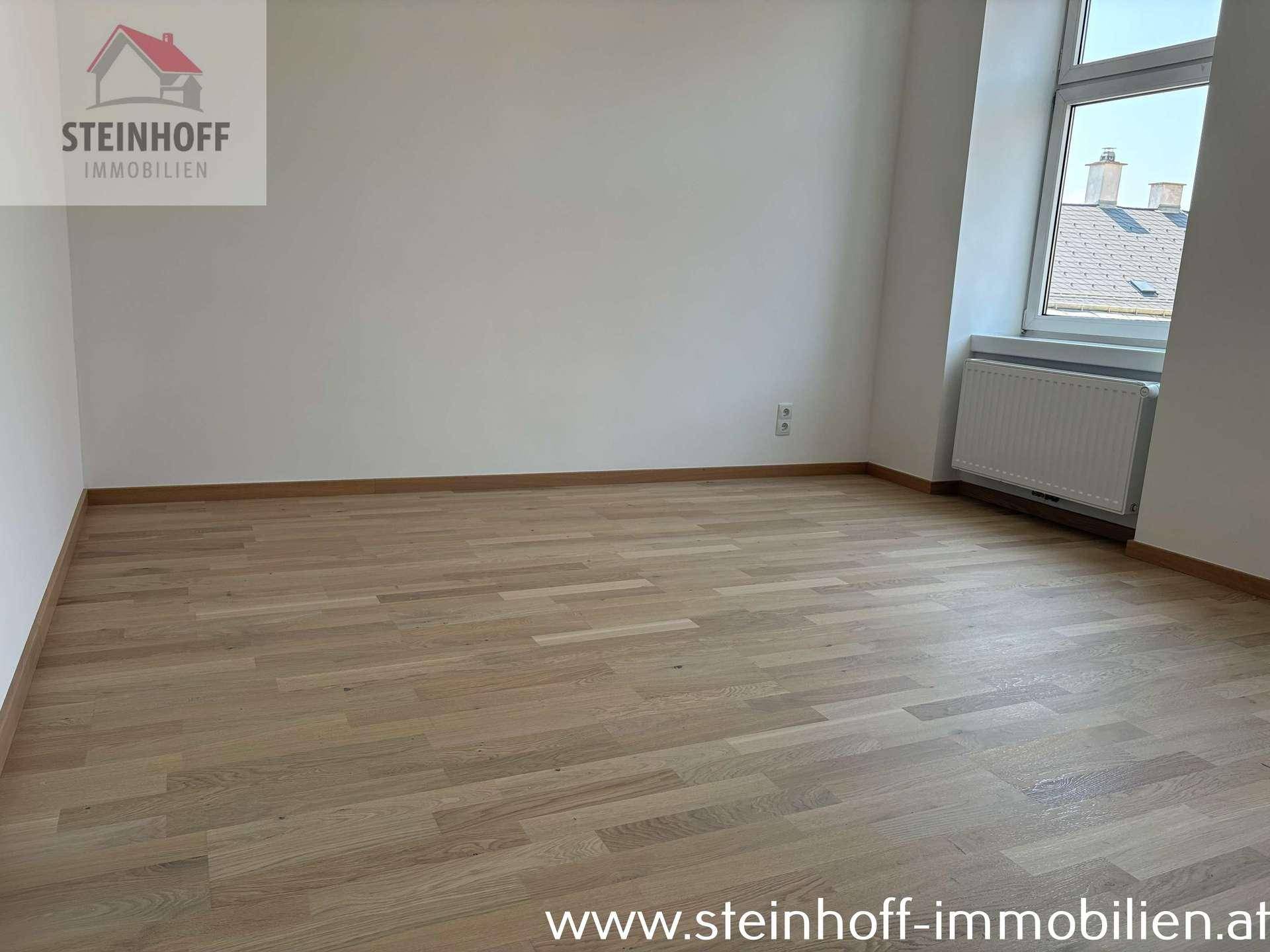 Helles Zimmer mit Parkettboden und einem Fenster, das einen Blick auf die Dächer der Stadt bietet.