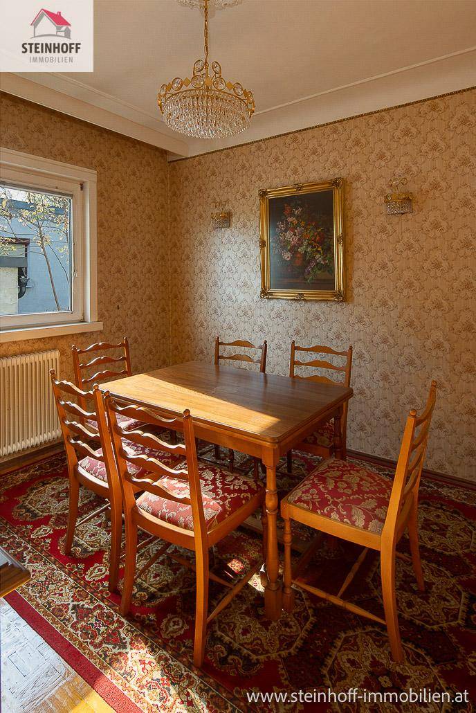 Esszimmer mit Holztisch, Stühlen und gemusterter Tapete, beleuchtet durch ein Fenster.