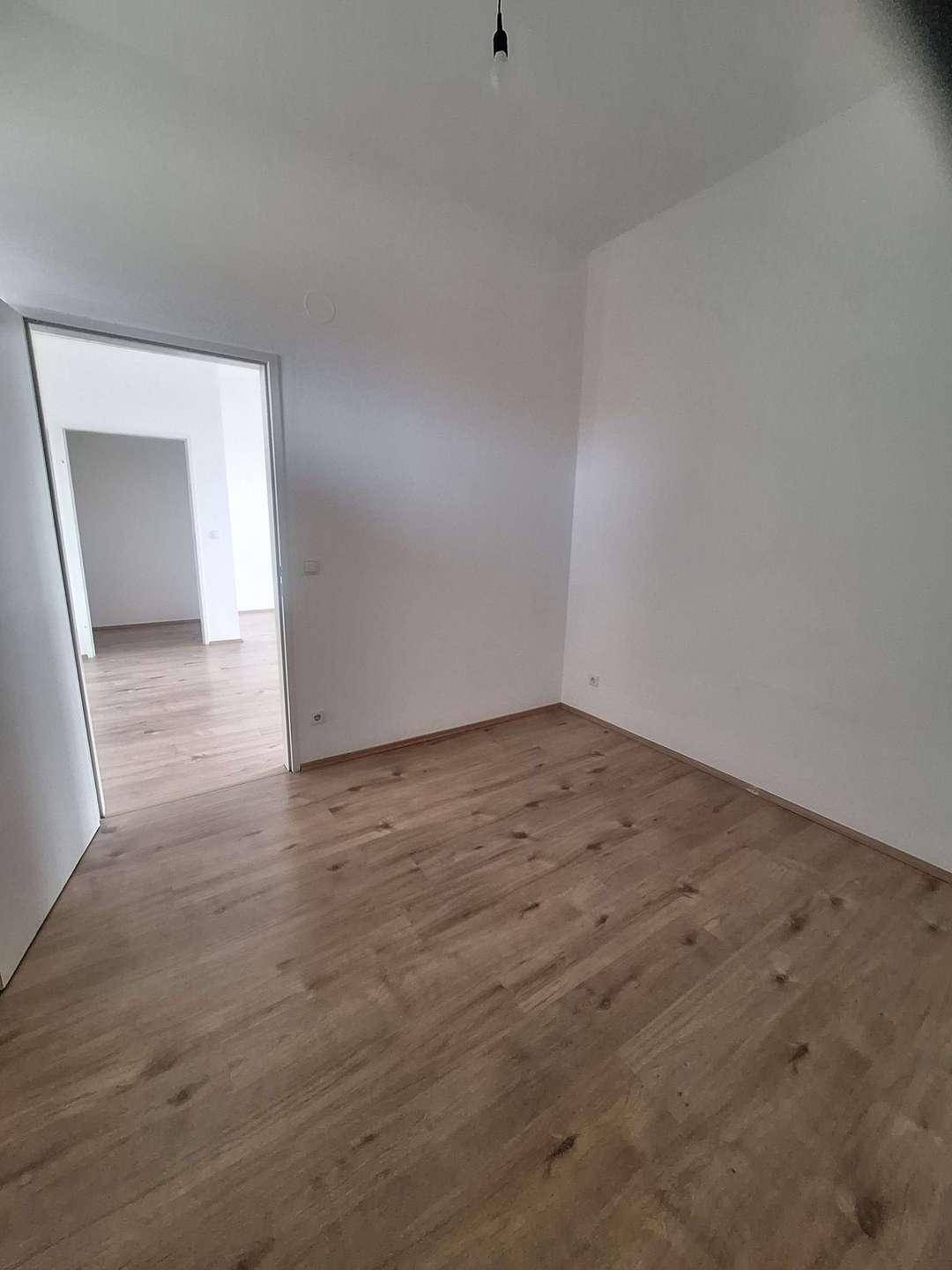 Helles, leeres Zimmer mit Laminatboden und weißen Wänden, eine offene Tür verbindet es mit einem weiteren Raum.