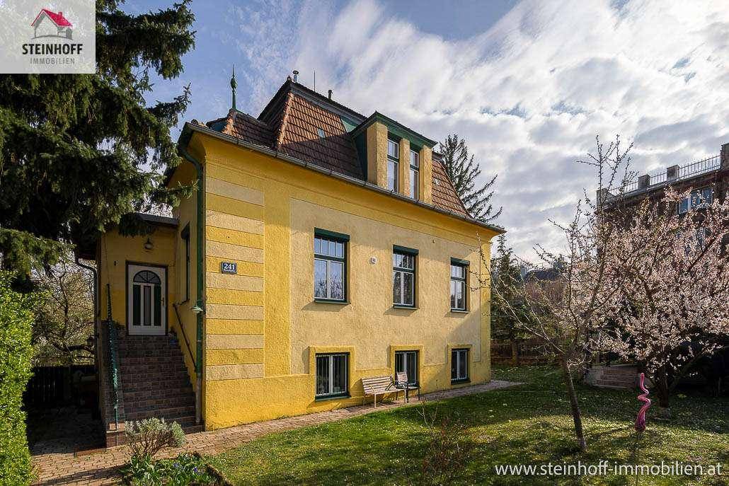 Charmantes gelbes Haus mit Mansarddach, grüner Umgebung und blühendem Baum.