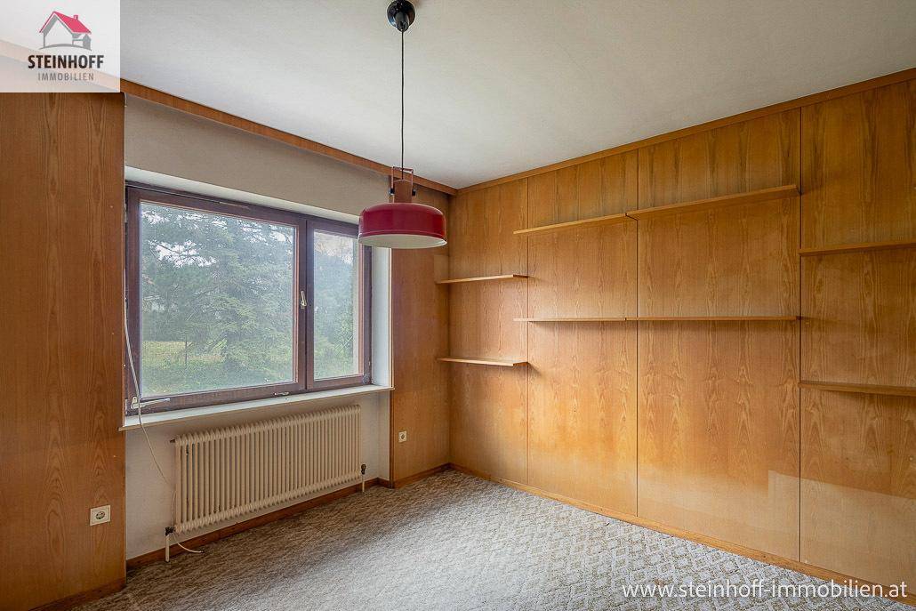 Zimmer mit Holzvertäfelung, Teppichboden und großem Fenster mit Blick ins Grüne.