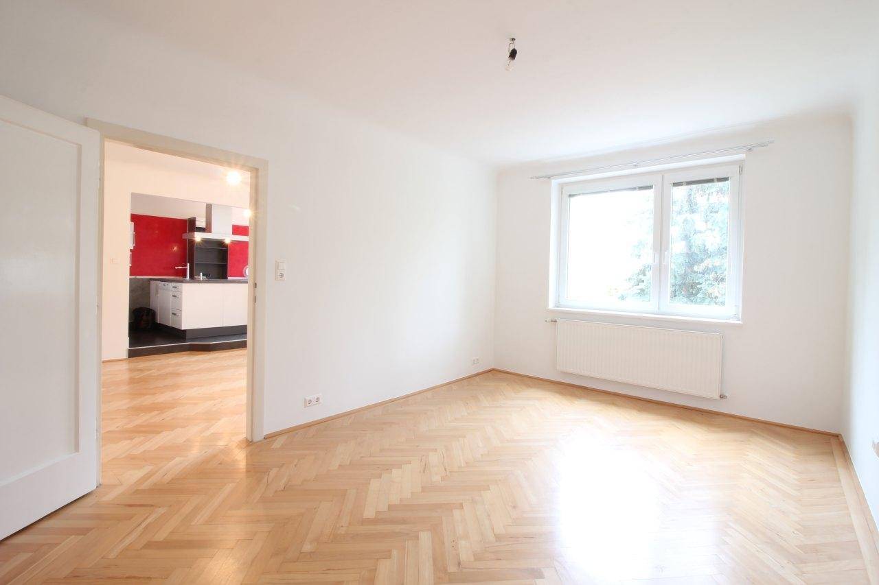 Geräumiger Wohnraum mit Fischgrätparkett, viel Tageslicht und Zugang zur Küche.