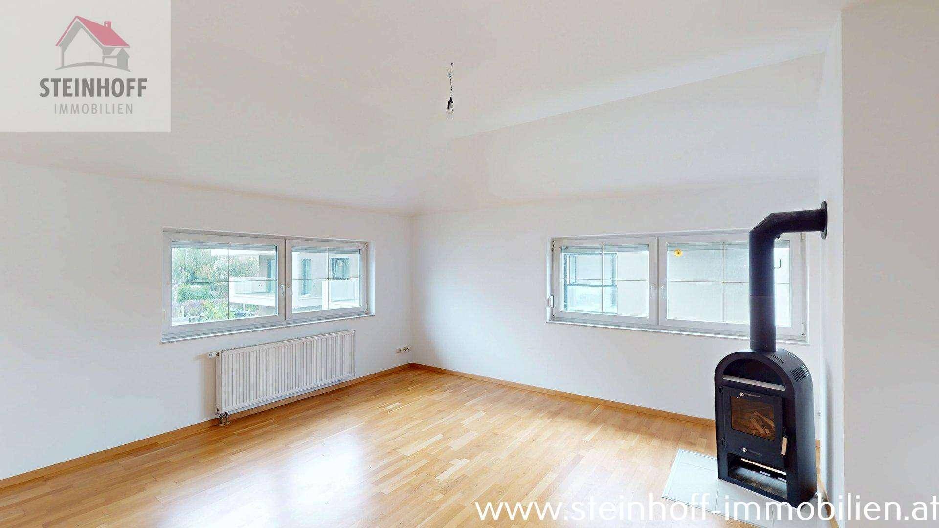Heller Raum mit Holzboden, zwei Fenstern und einem Kaminofen, ideal für einen Wohnbereich.