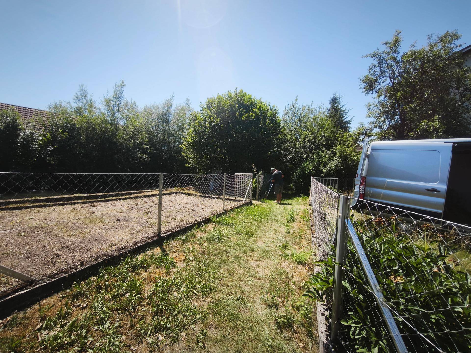Gartenbereich mit Zaun und einem Transporter im Hintergrund, umgeben von Bäumen.
