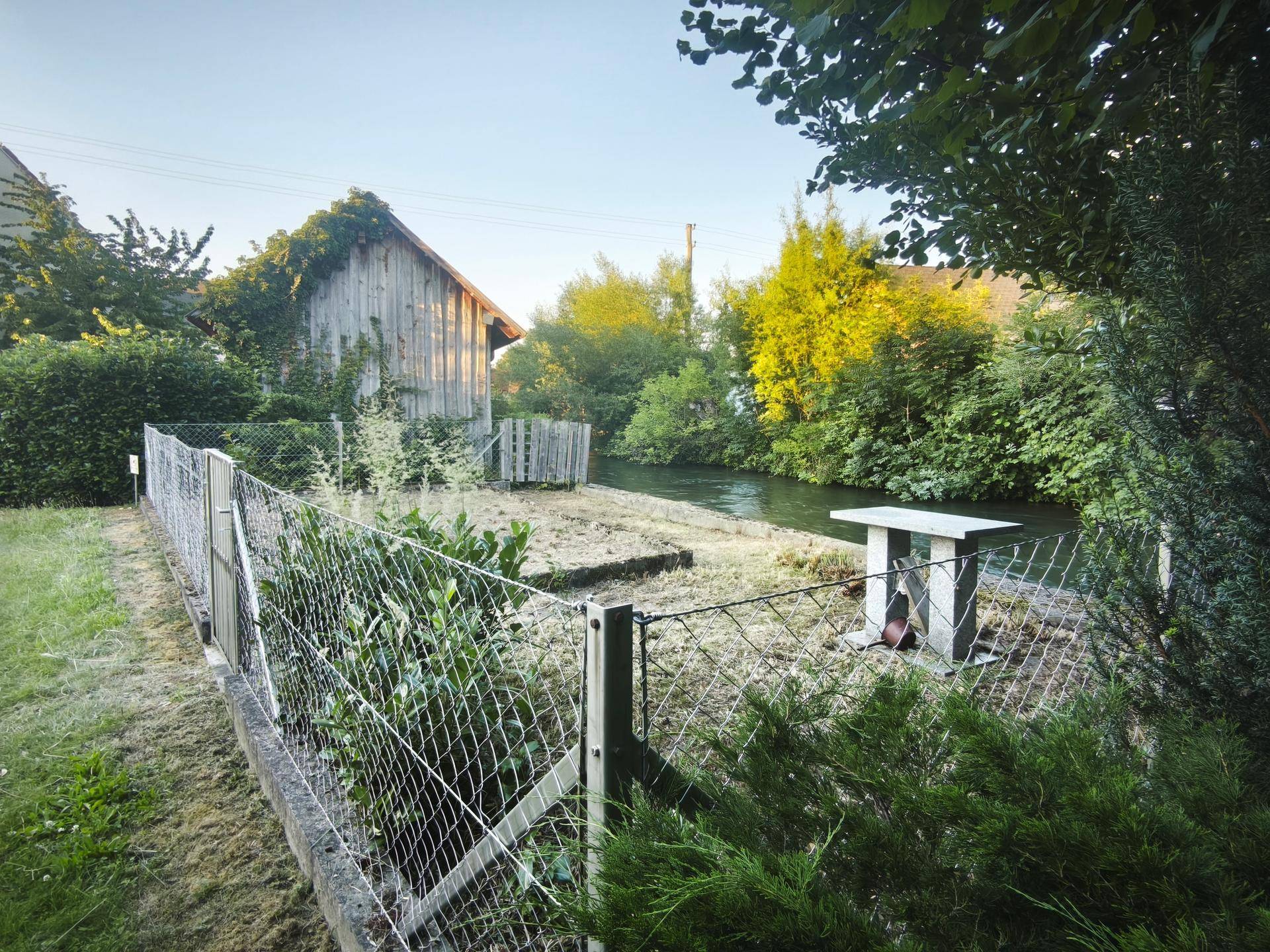 Eingezäunter Gartenbereich mit einem Steintisch und Blick auf den Fluss und den Holzschuppen.