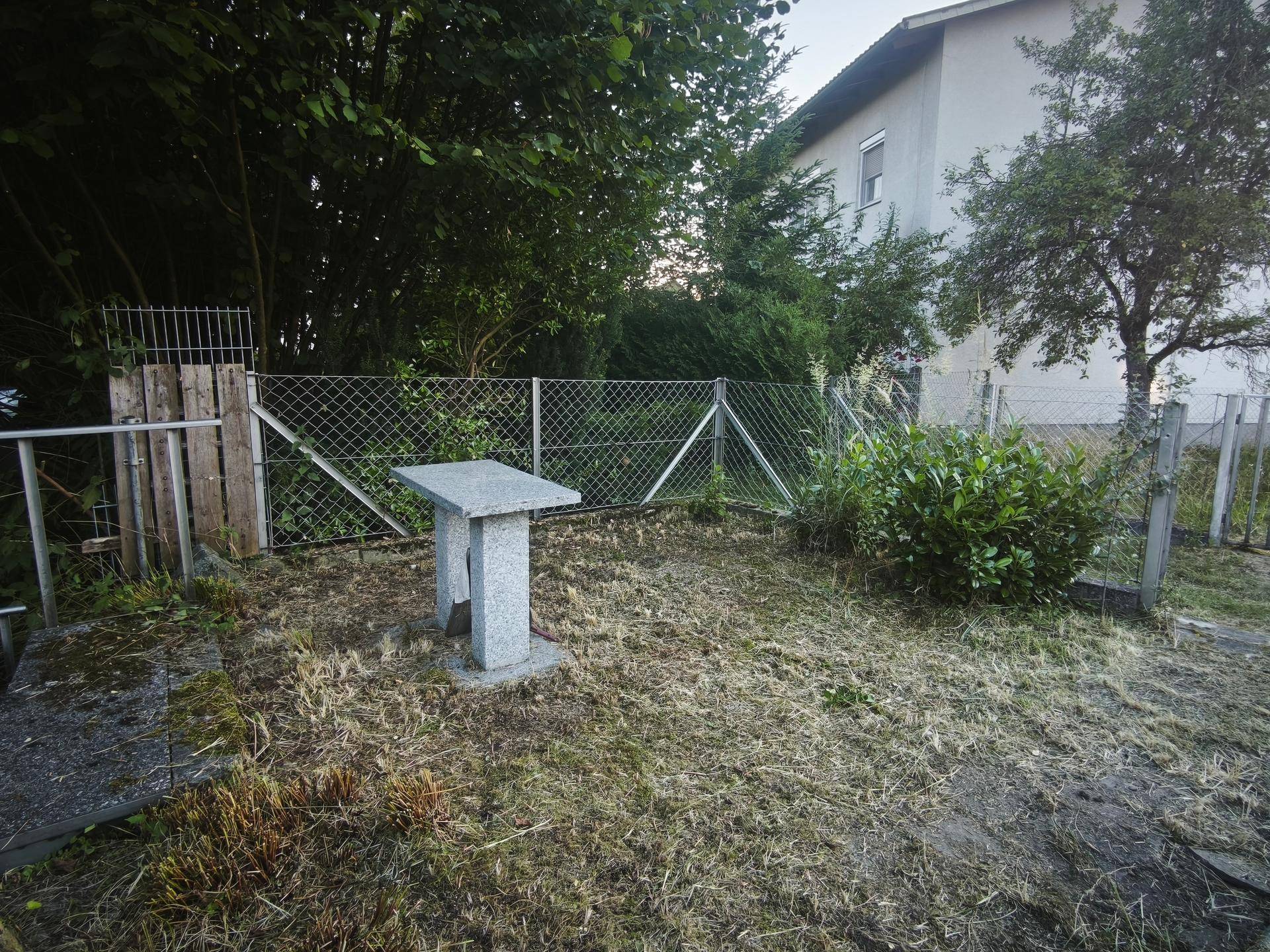 Gartenbereich mit Rasen, Bäumen und einem Maschendrahtzaun, angrenzend an ein Wohngebäude.