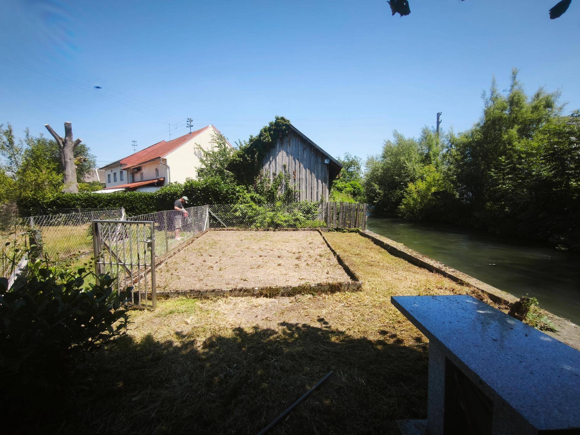 Sonniges Gartengrundstück am Fluss mit einem Steintisch und einem alten Holzschuppen.